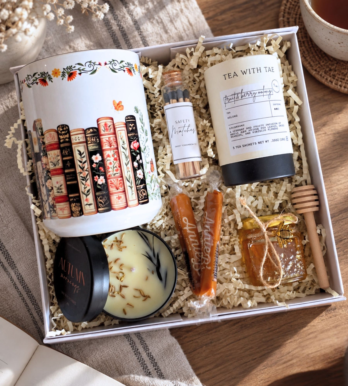 Overhead view of the open gift box, showing the mug, tea tin, honey jar, candle, matches, and artisan caramels nestled in crinkle fill in a white magnetic gift box