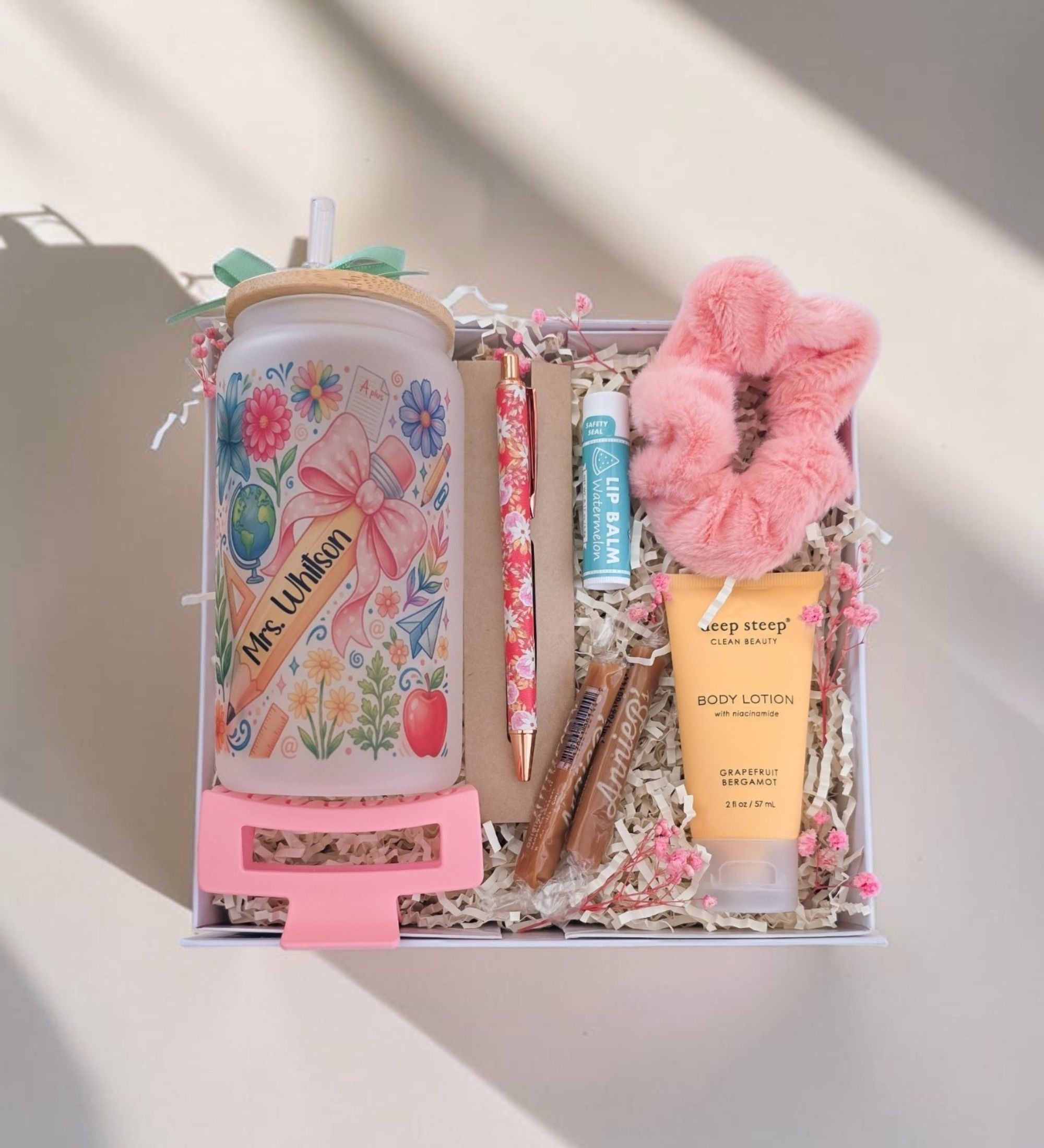 Overhead view of open Deluxe teacher gift box with personalized Mrs. Whitson coquette floral tumbler, watermelon lip balm, Grapefruit Bergamot body lotion, floral pen, artisan caramels, pink scrunchie, pink hair clip in white crinkle paper with dried pink florals