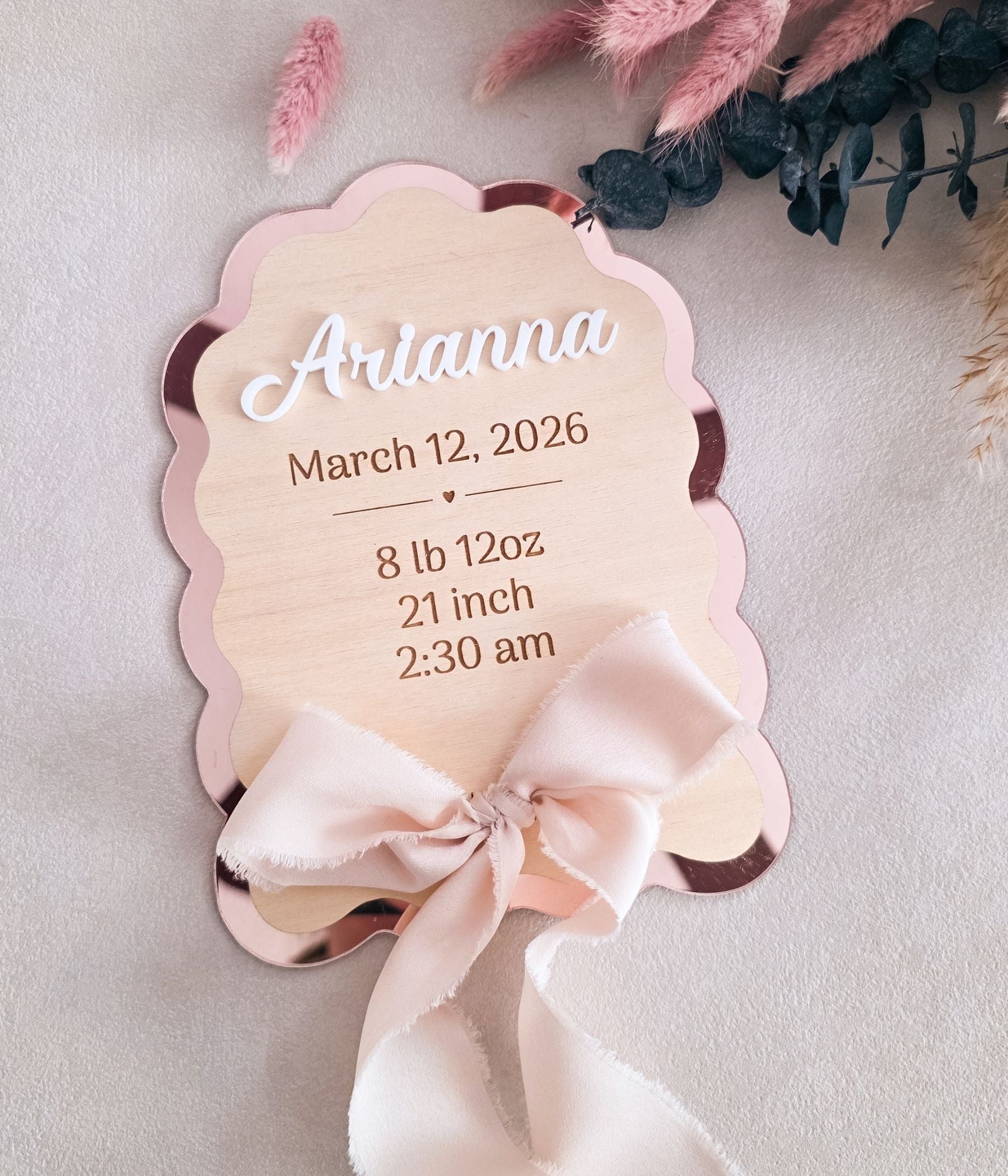 Personalized baby birth stats sign in rose mirror acrylic and birch wood, scalloped edges, engraved with name Arianna, birth date, weight, length, and time — flat lay with pink dried florals and chiffon ribbon bow