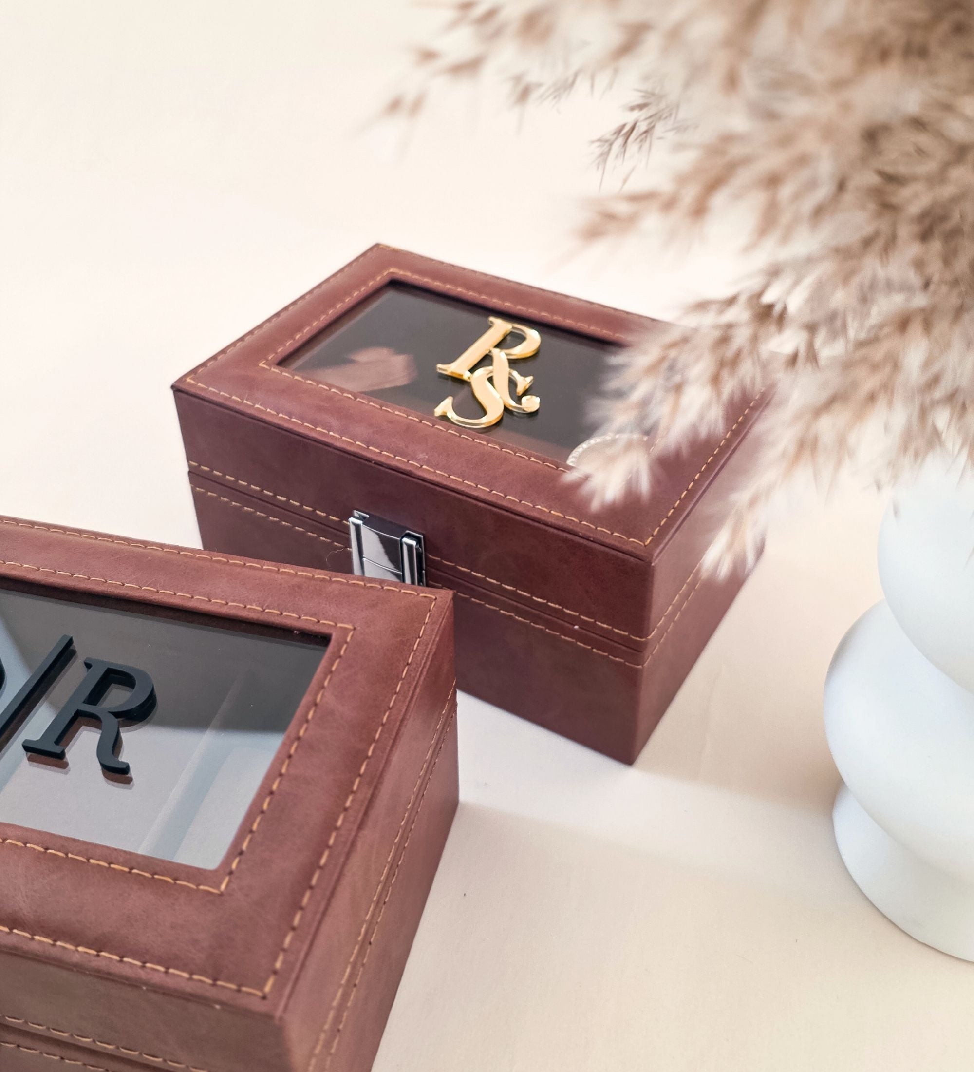Two cognac leatherette watch boxes together — one closed with "KD" black acrylic monogram, one open showing three padded watch pillow compartments, displayed with white ceramic vase