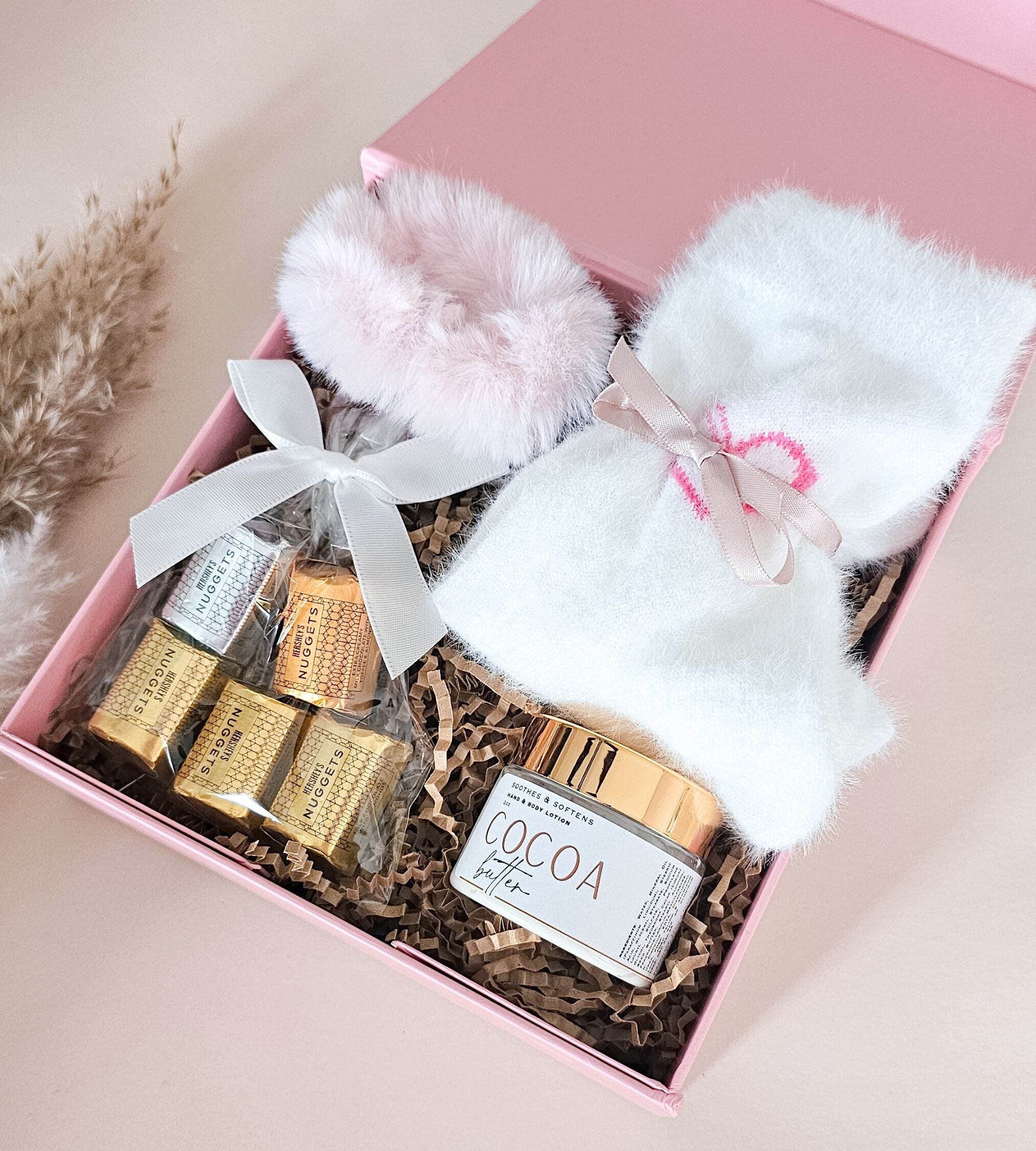 A pink gift box filled with a white fluffy socks tied with a pink ribbon, a pale pink fluffy hairscrunchie, a jar of cocoa butter, and wrapped chocolates on shredded paper.