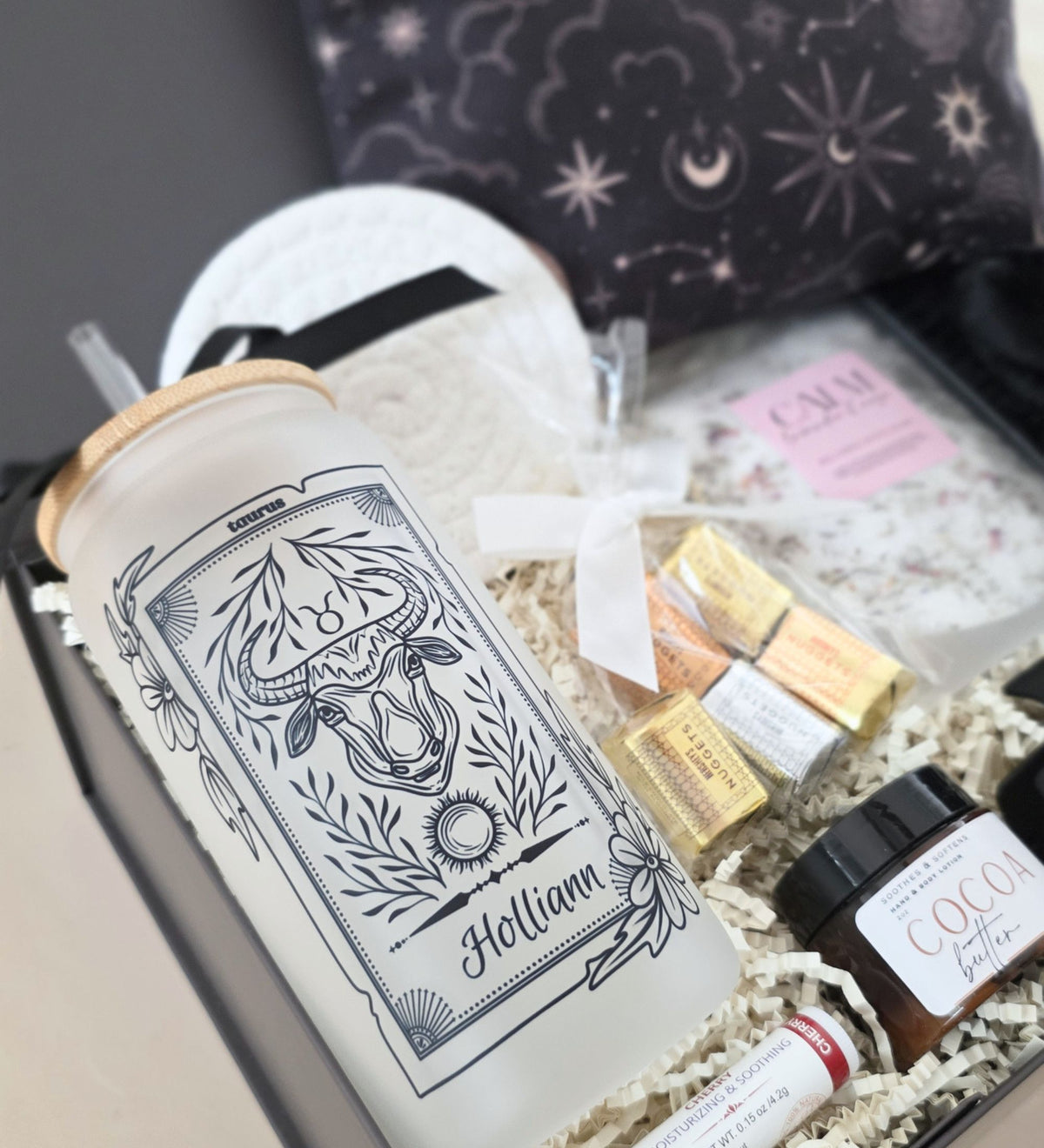 Styled overhead view of open Ultimate Zodiac Box with celestial zipper pouch, Taurus tumbler "Holliann," bath soak, spa headband, hair clip, lotion, nuggets, and lip balm