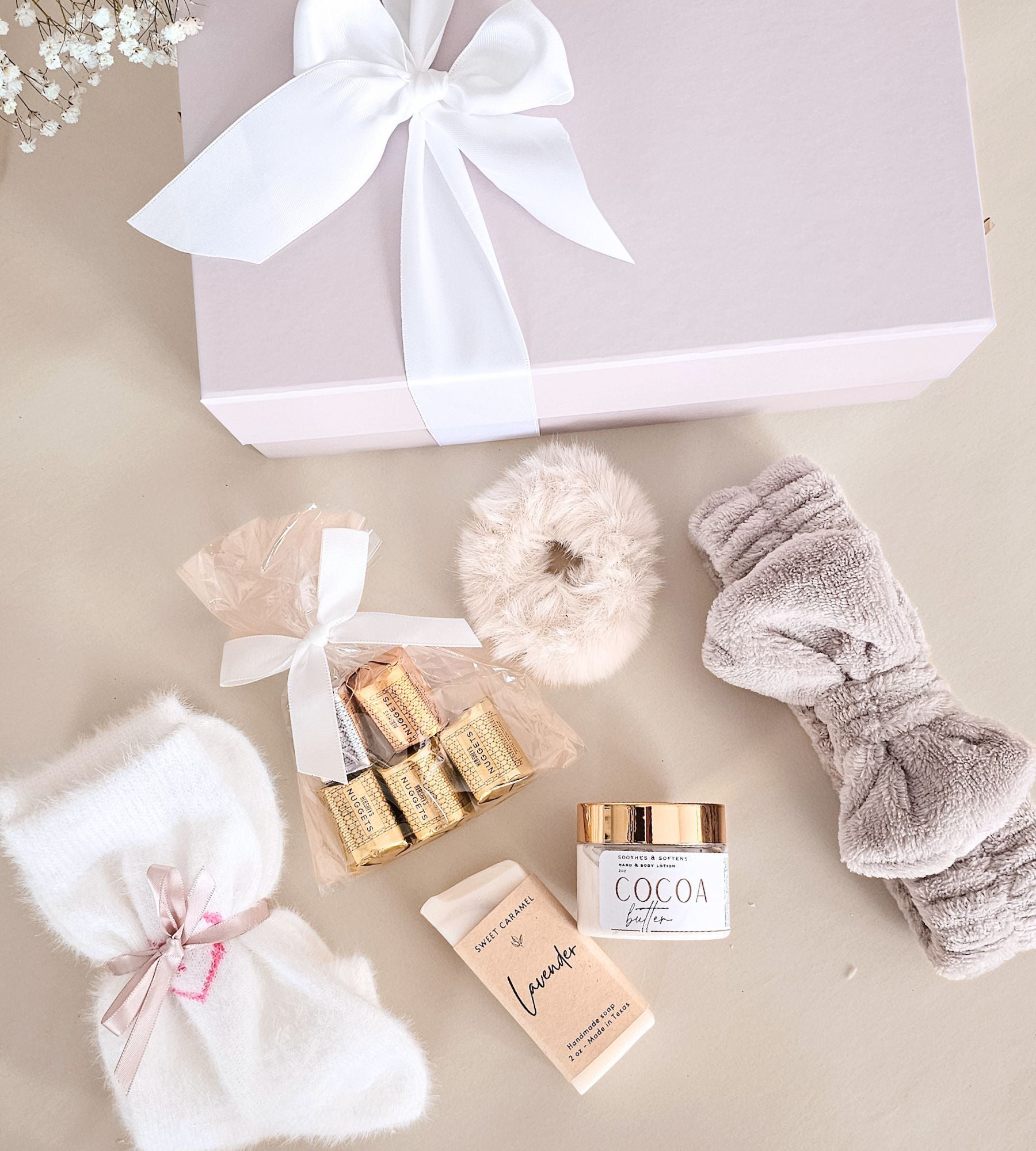 A luxury gift set arranged on a neutral beige surface, featuring a pale beige gift box with a white ribbon, a white plush socks, a gray plush headband, a small bag of gold-wrapped chocolates, a jar of cocoa butter lotion, and a handmade soap bar