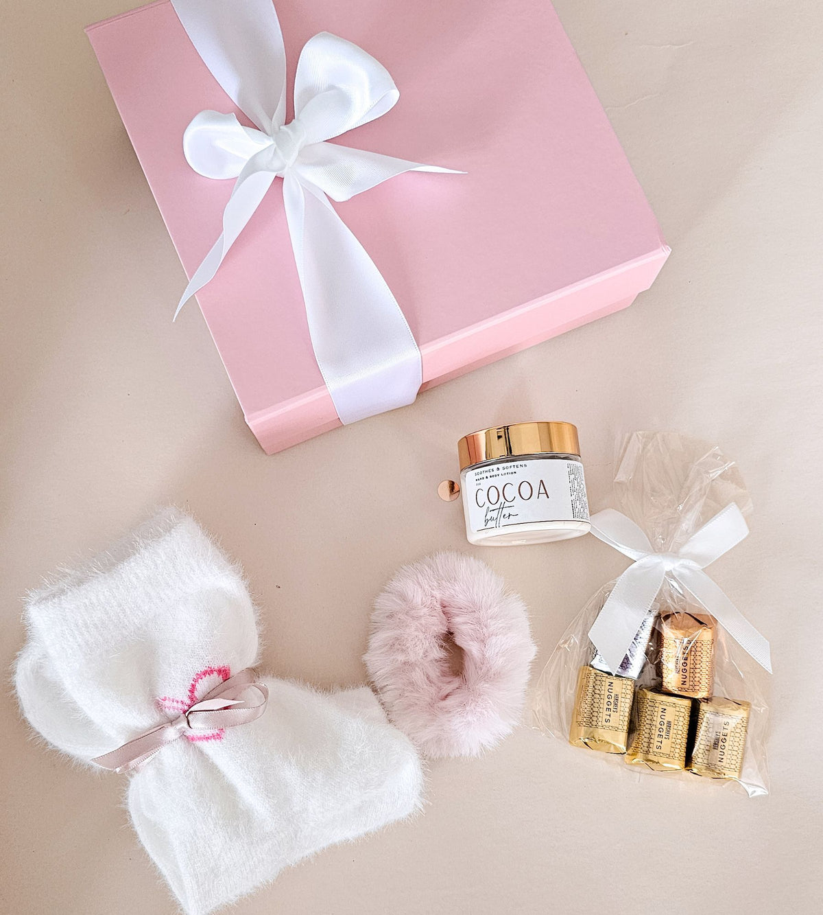 A pink gift box tied with a white satin bow, arranged on a beige surface with a white fuzzy socks, a pink scrunchie, a small lotion jar labeled COCOA, and wrapped chocolates tied in a clear bag with a white ribbon.