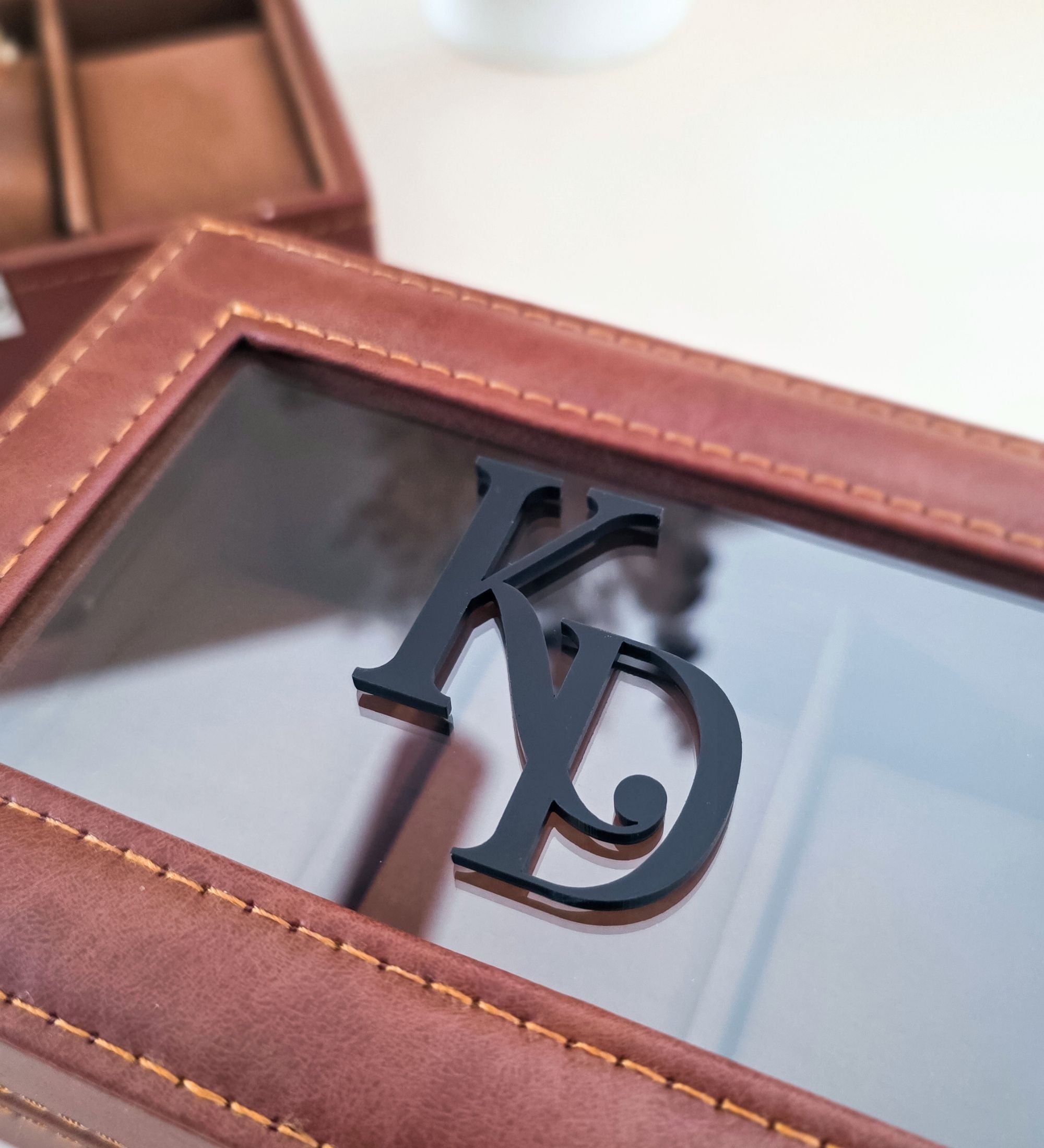 Close-up of cognac leatherette watch box with black 3D acrylic monogram, showing polished silver latch detail and contrast stitching