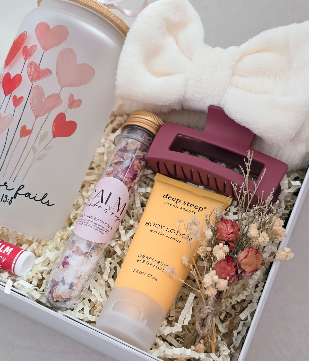 A curated gift box set containing a frosted glass canister with a heart pattern, a tube of deep steep body lotion, a jar of bath salts, a white plush bow, and a dusty rose hair claw clip, arranged in white paper shred.