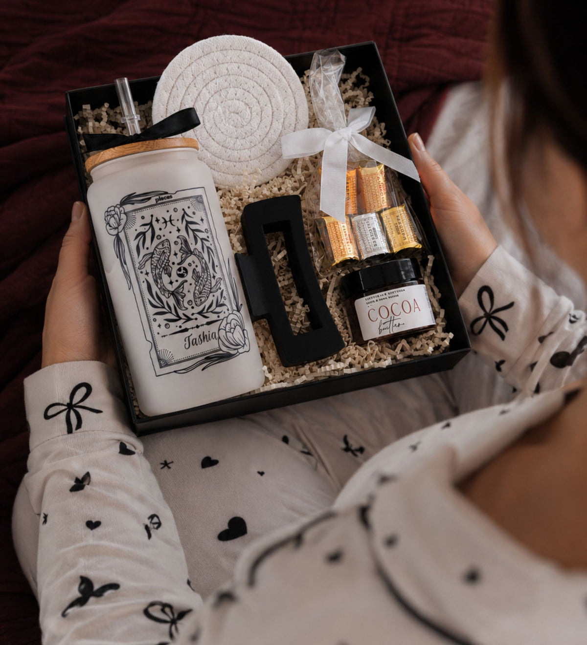 A gift set in a black box held by a person wearing a white pajama top with black bow and heart prints. The set includes a white tumbler with a black illustration and straw, a white woven coaster, a black letter 'F', gold and white wrapped chocolates, and a small jar labeled 'COCON'.