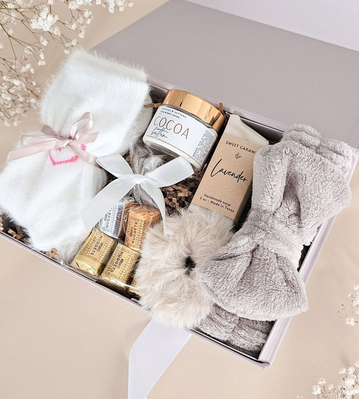 A curated spa gift box filled with self-care items, including a white plush bath towel tied with a pink ribbon, a jar of cocoa butter, a boxed lavender hand cream, gold-wrapped chocolates, and a fluffy cream bath mat, arranged in a light beige box on a beige surface.