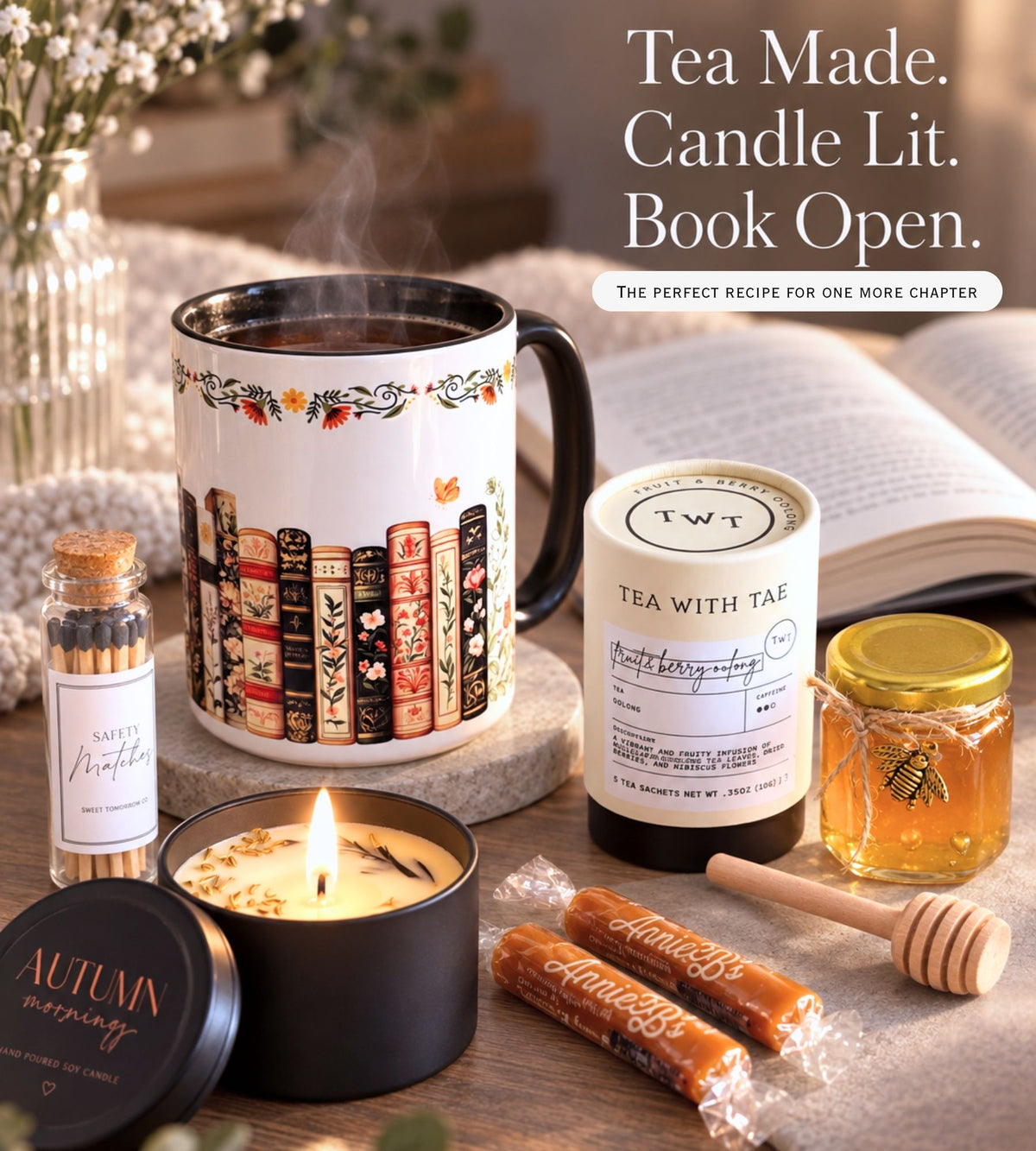 A cozy autumn-themed bookish candle and tea gift set arranged on a wooden surface with an open book in the background. The set includes a white ceramic mug printed with colorful book spines, a black candle tin labeled 'Autumn mornings,' wrapped caramel candy sticks, a small bottle of matches labeled 'Safety matches,' a jar of honey, a wooden honey dipper, and a tea jar with a gold lid labeled 'Tea with Tae.