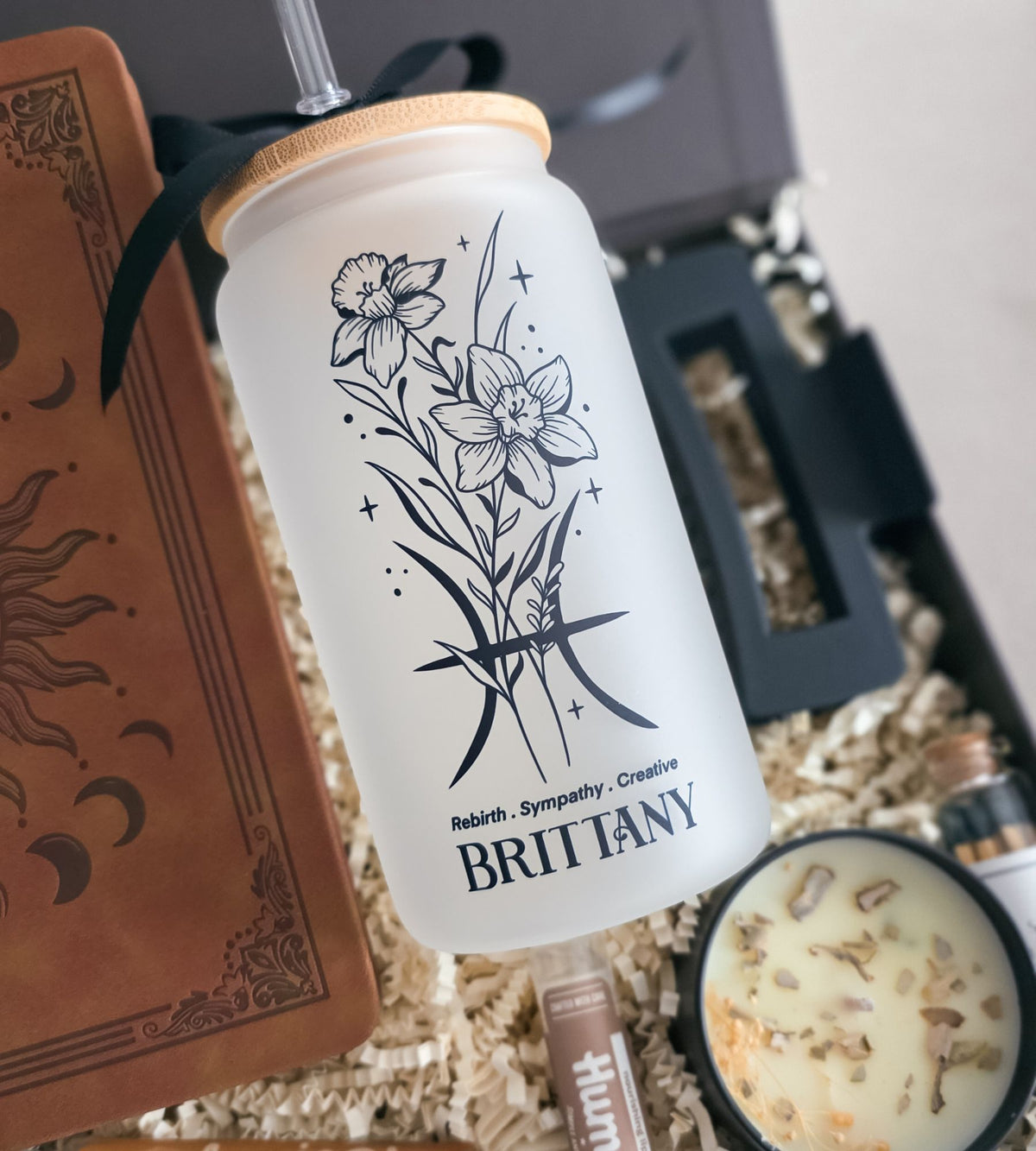 A frosted glass tumbler with a bamboo lid and black straw, featuring a botanical line art design and the name 'BRITTANY' on the front. The tumbler is styled in a gift box with a brown leather journal, a candle, and other gift items.