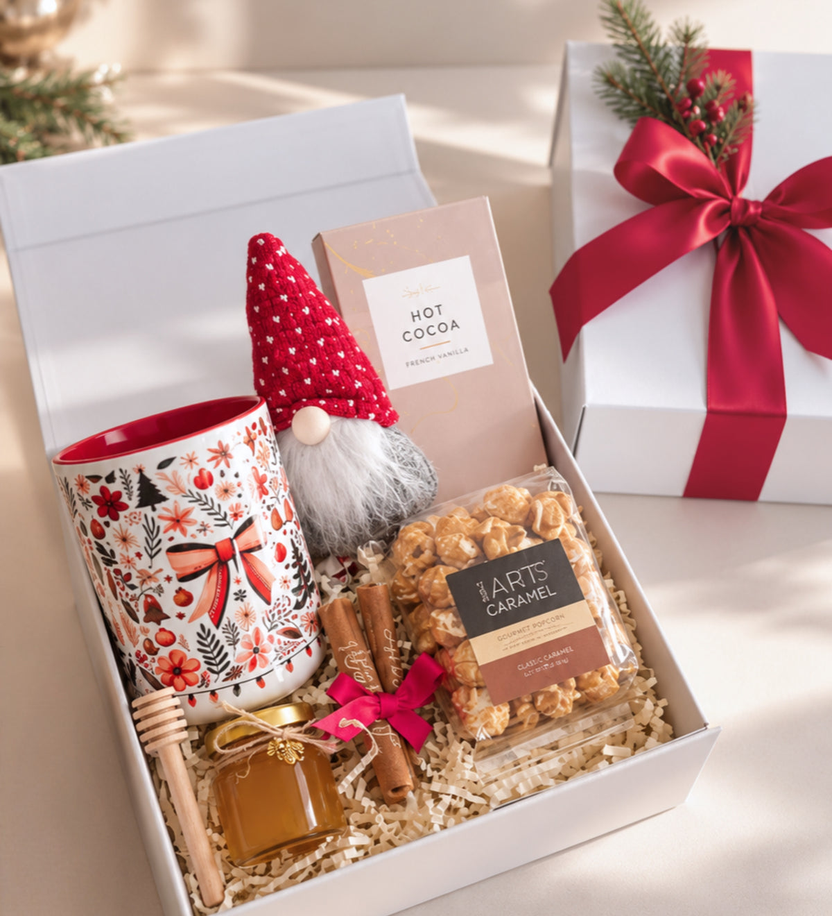 Open white magnetic gift box with a red-patterned holiday mug, soft gnome, hot cocoa, caramel popcorn, honey and caramels, beside a ribbon-tied box