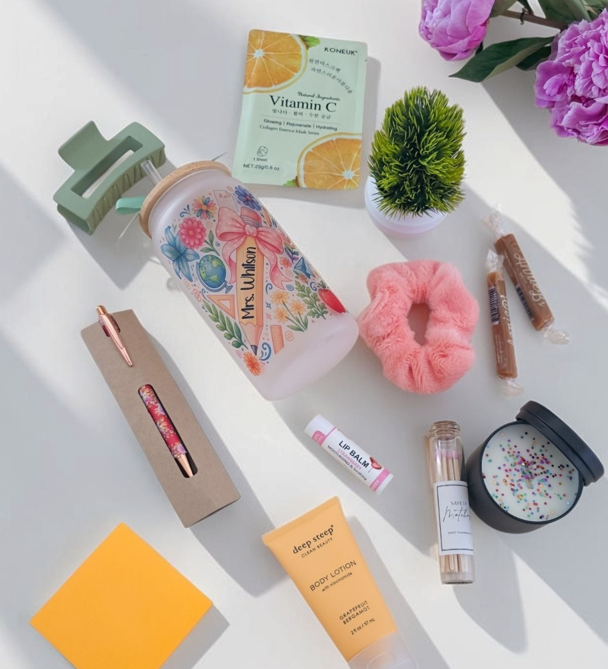 Angled lifestyle view of open teacher gift box showing personalized coquette floral tumbler with green bamboo lid, floral pen, watermelon lip balm, Grapefruit Bergamot body lotion, artisan caramels, pink scrunchie, pink hair clip in white crinkle paper with pink peony styling

