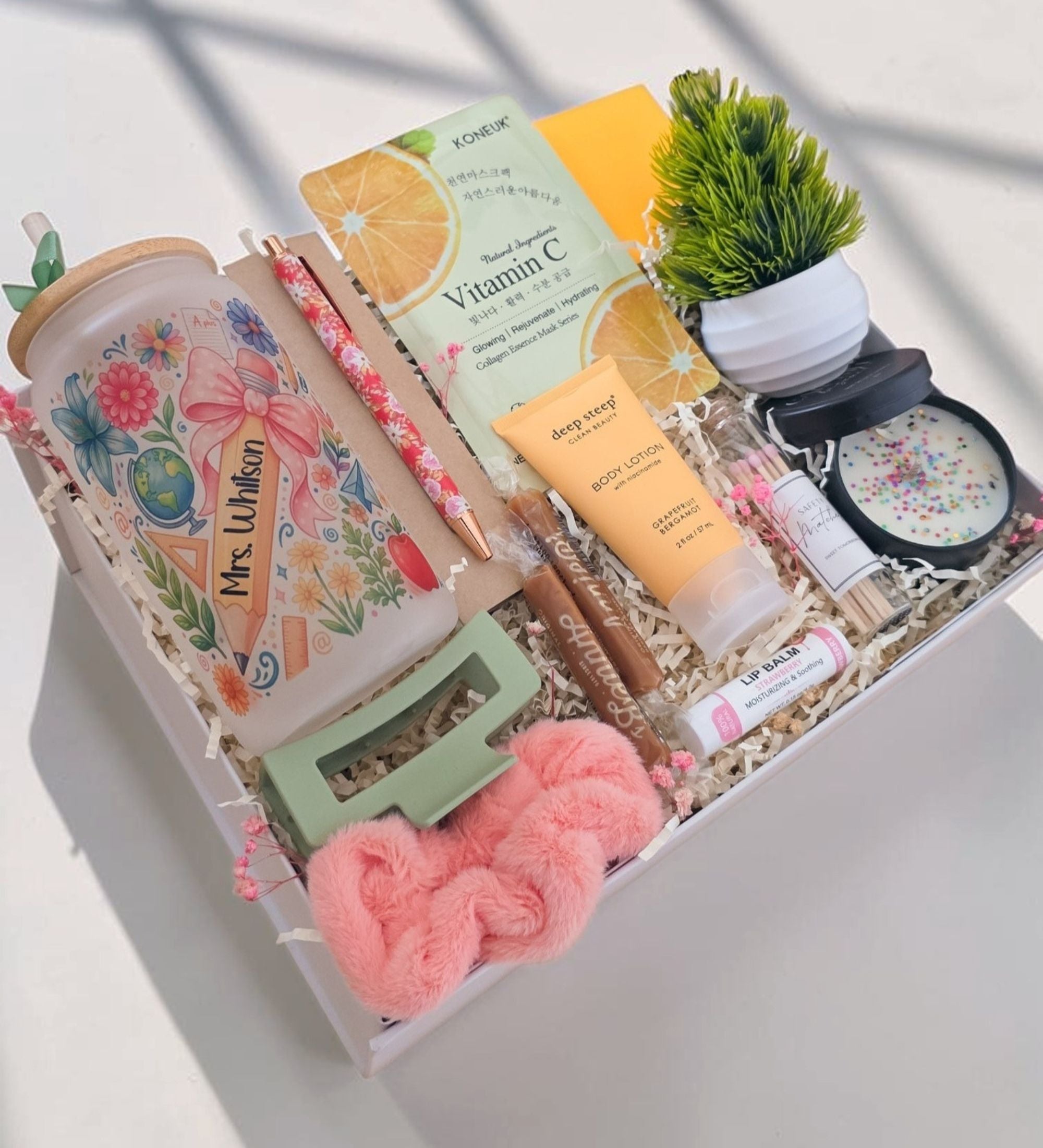 Overhead angled view of open Ultimate teacher appreciation gift box in white crinkle paper showing personalized coquette floral tumbler, Vitamin C collagen face mask, Grapefruit Bergamot body lotion, artisan caramels, sprinkle soy candle, miniature faux desk plant, apothecary safety matches, floral pen, pink scrunchie, green hair clip