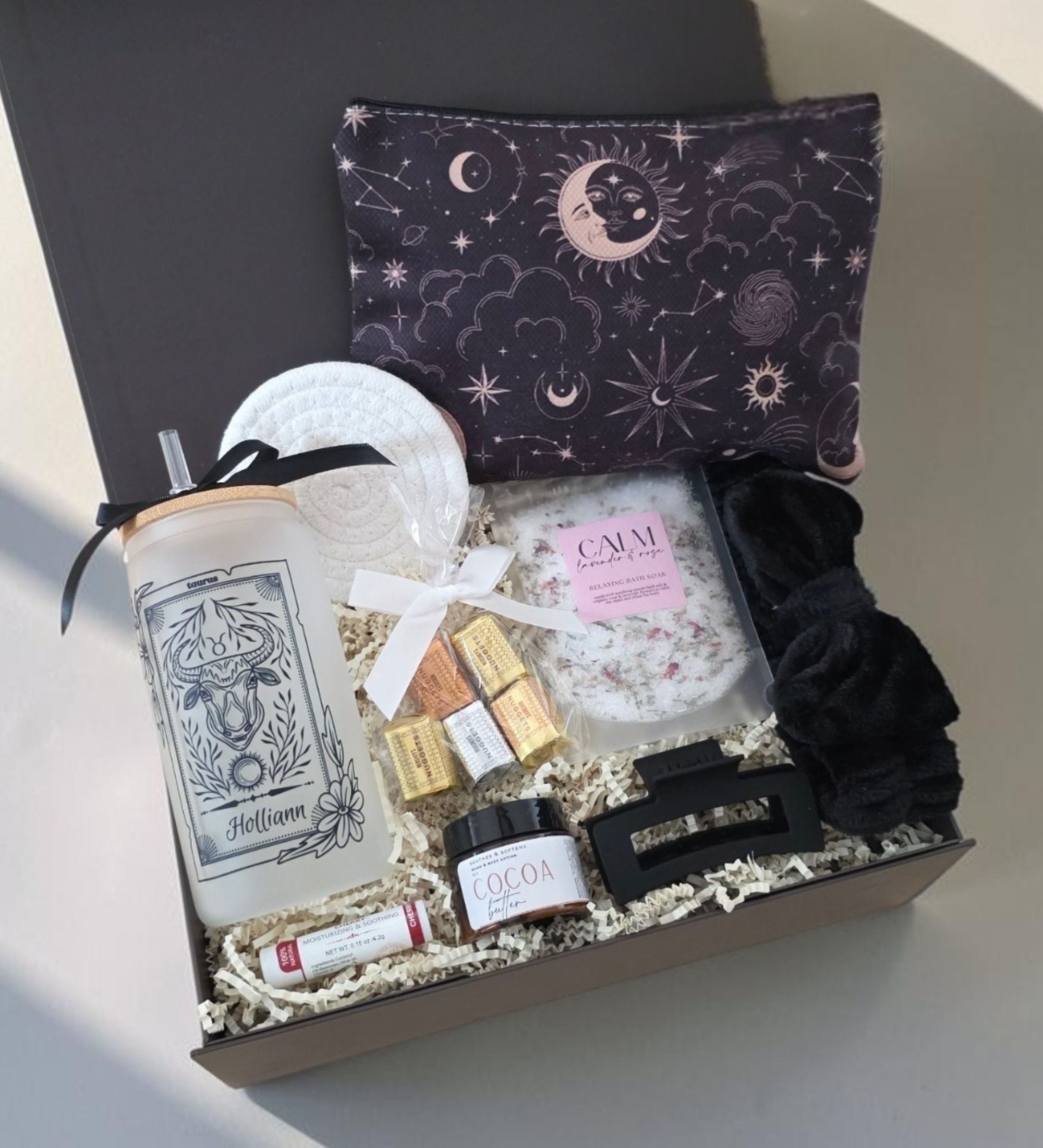 Open black gift box showing full Ultimate Zodiac Box with Taurus tumbler "Holliann," celestial black zipper pouch, bath soak, spa headband, matte hair clip, chocolate nuggets, cocoa butter lotion, lip balm