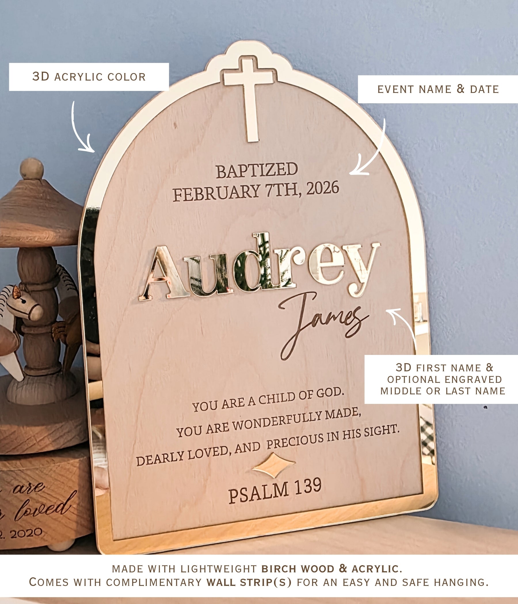 Personalized arched wooden baby milestone plaque with a cross at the top and custom text including 'Audrey James' and 'Baptized February 7th, 2026,' shown hanging against a blue wall with decorative objects beside it. Personalized christening sign displayed upright, showing the full layout of name, date, and verse