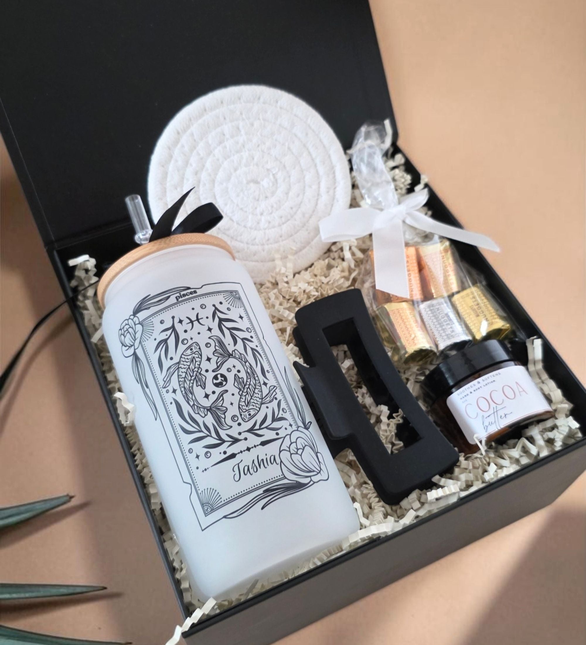 Open black magnetic gift box showing Deluxe Zodiac Box contents: personalized Pisces frosted glass tumbler with "Tashia" name, matte black hair clip, drink coaster, chocolate nuggets, cocoa butter lotion in white crinkle paper