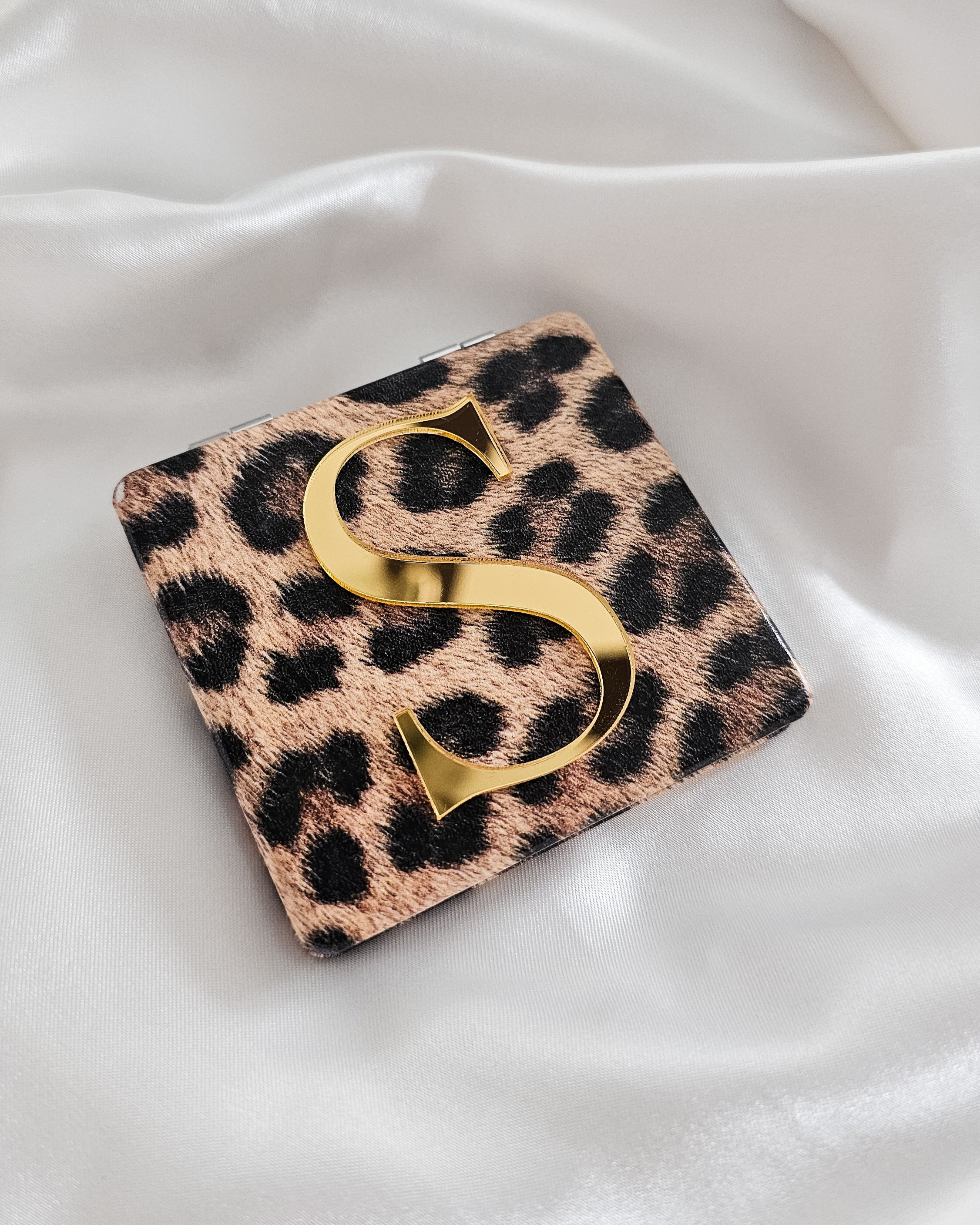 Personalized leopard-print compact mirror with a bold gold 3D letter "S" on top, displayed on white satin fabric.