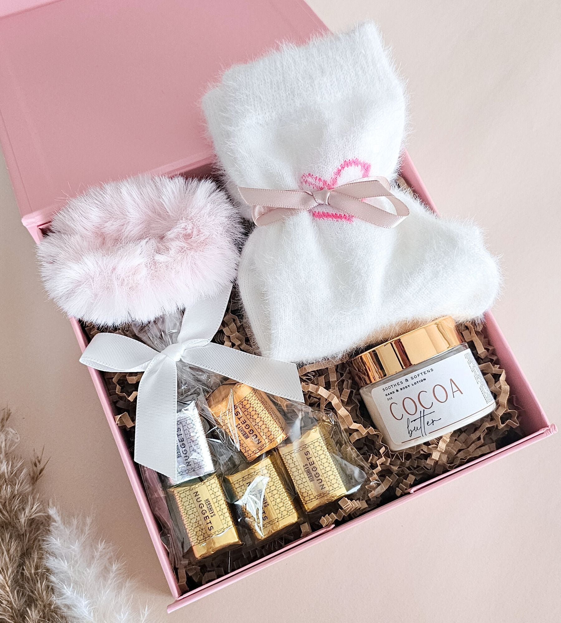 A pink gift box filled with spa treats and self-care items, including a white fuzzy socks tied with a pink ribbon, a fluffy pink scrunchie, wrapped chocolates, and a lotion jar labeled COCOA.