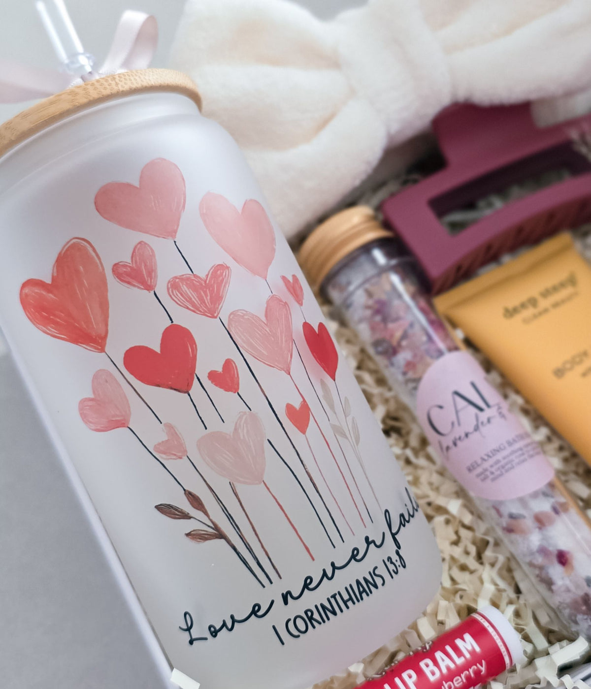 A frosted glass tumbler with a bamboo lid and straw, decorated with pink and red heart illustrations and the text 'Love never fails I CORINTHIANS 16:14', shown in a gift basket with bath items and self care items