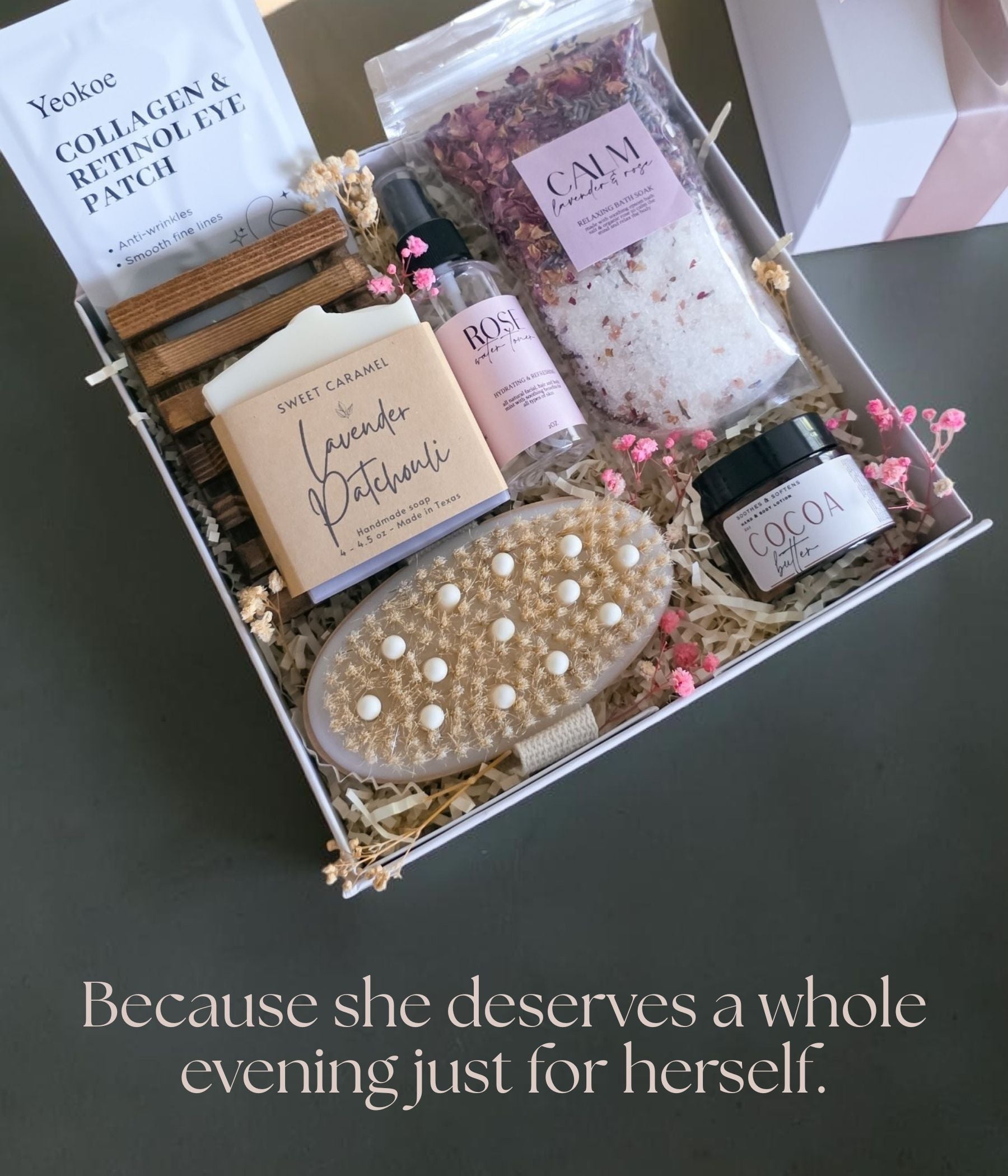 A curated self-care gift box filled with spa items, including a collagen and retinol eye patch, rose body wash, lavender patchouli soap, bath salts, a scrubber, and a cocoa butter, arranged in a white box with shredded paper filler. A text overlay at the bottom reads, 'Because she deserves a whole evening just for herself.