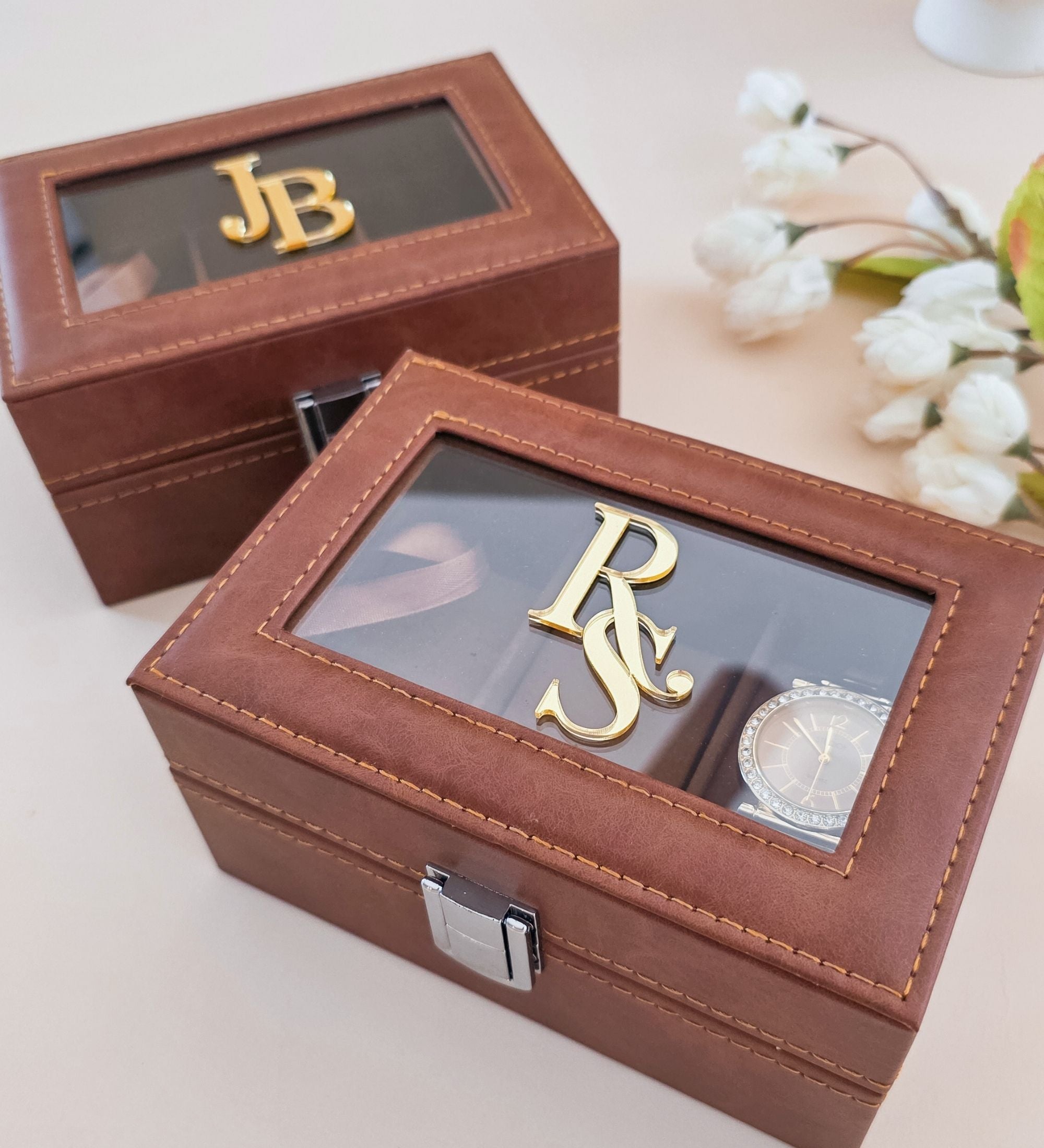 Two cognac leatherette watch boxes side by side, one small with gold monogram "JB" in Style A and one flat-style with gold "RS" monogram and watch visible inside, surrounded by white florals