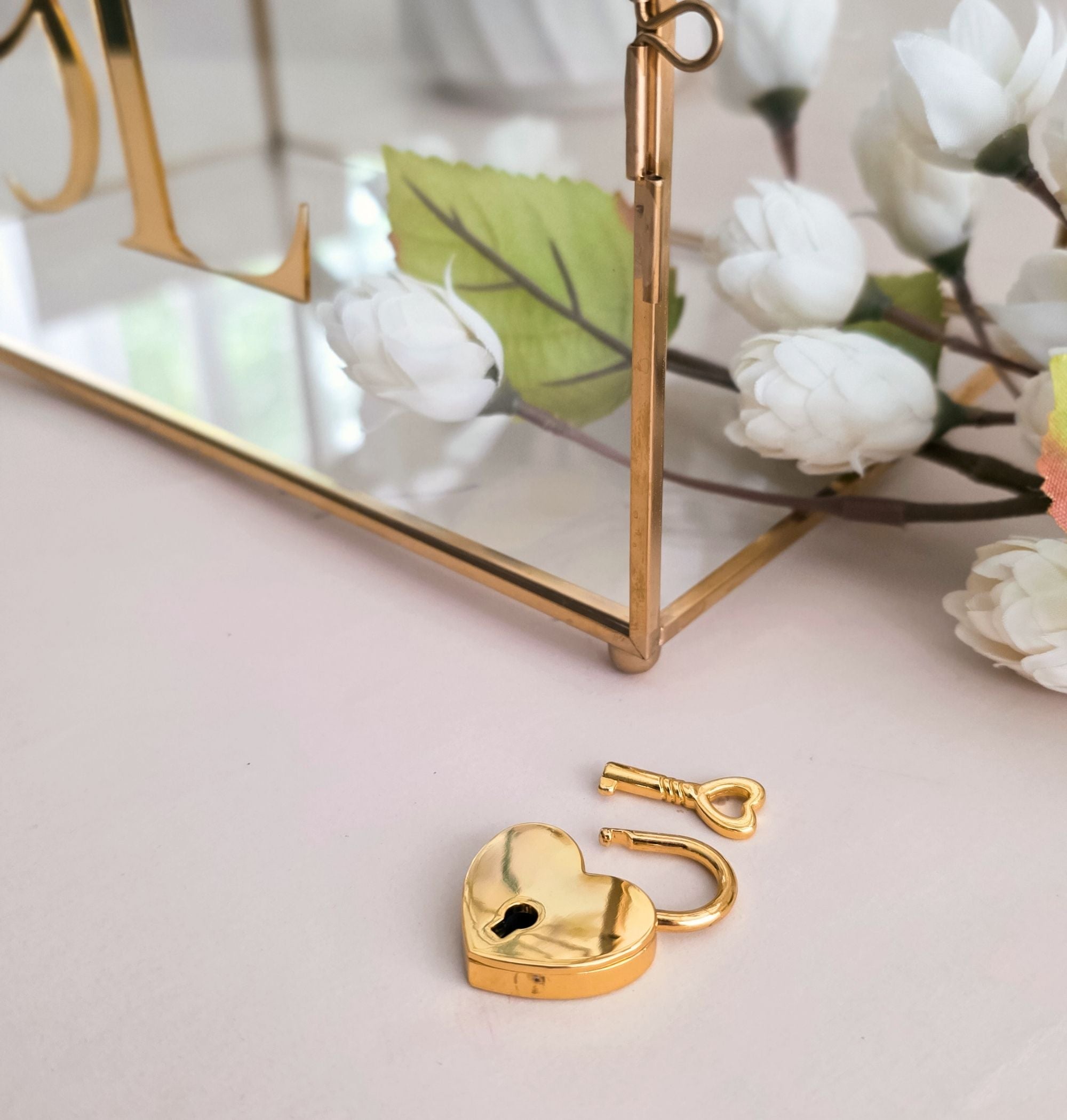 Close-up of gold heart-shaped padlock and key included with the personalized glass wedding card box, displayed beside white flower blossoms