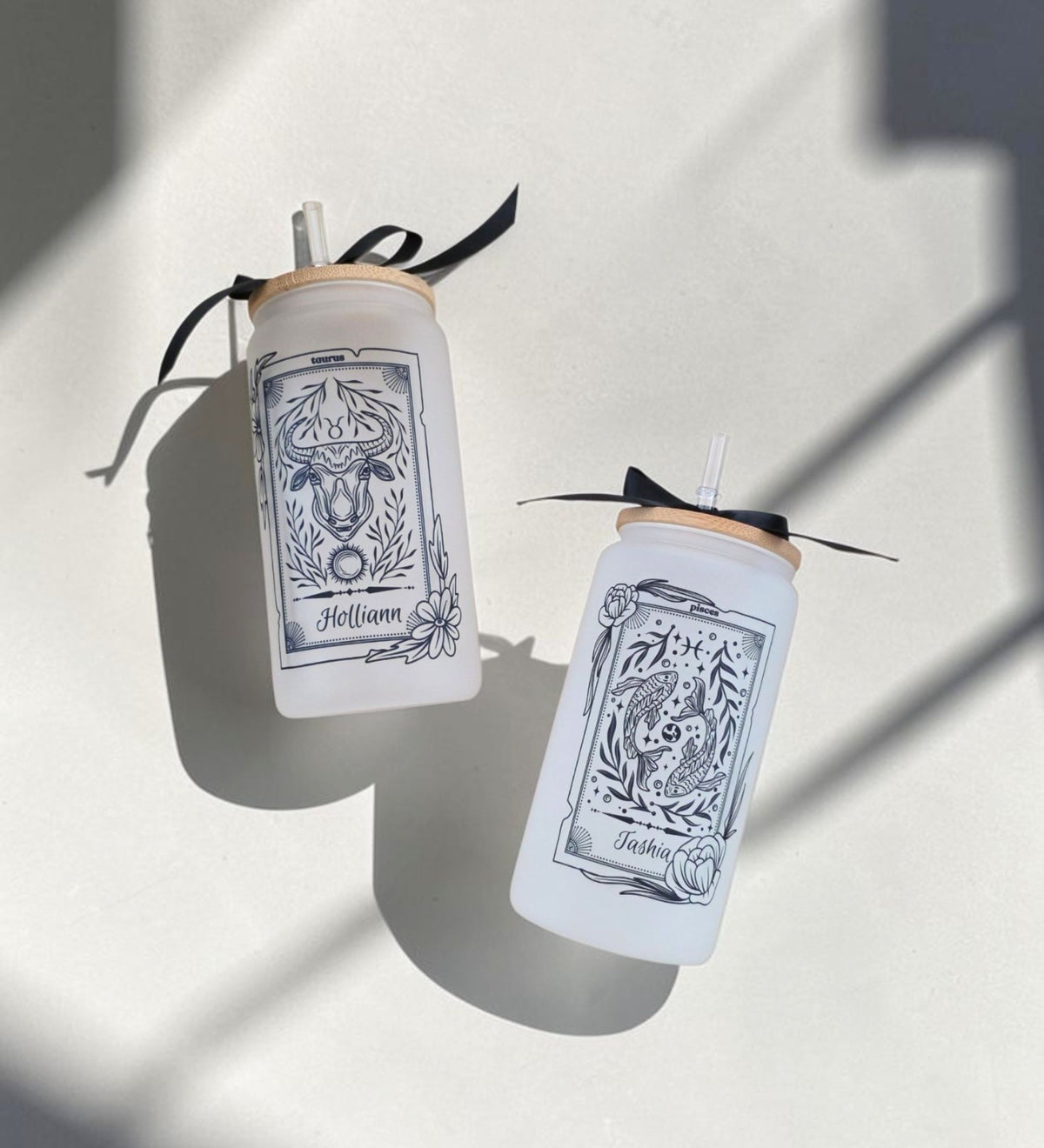 Two personalized frosted glass zodiac tumblers side by side — Taurus "Holliann" and Pisces "Tashia" — bamboo lids with black satin ribbon bows, dramatic shadow styling