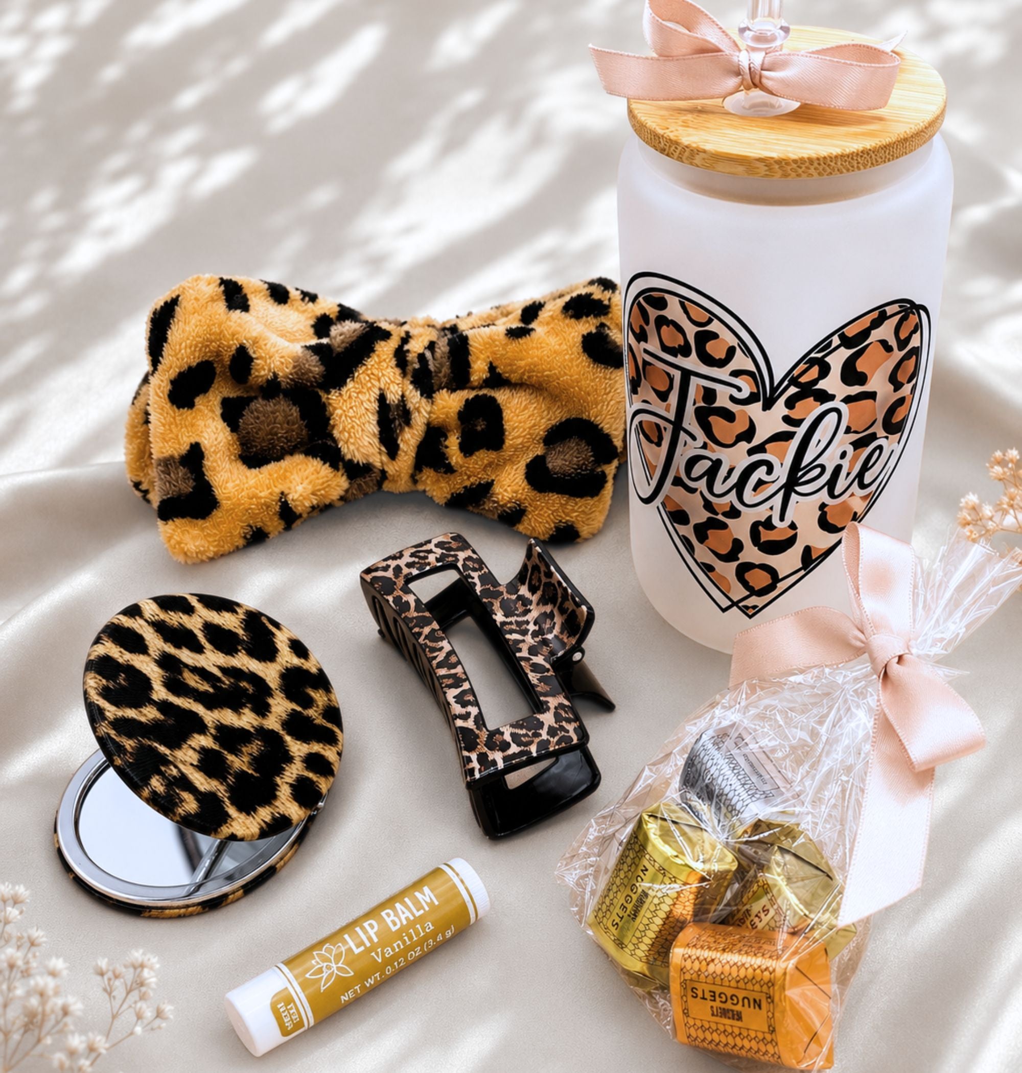 Flat lay of all Cheetah Obsessed gift box contents on white fabric: personalized frosted glass leopard tumbler with bamboo lid, leopard spa bow headband, leopard compact mirror open, leopard hair clip, vanilla lip balm, Hershey's Nuggets with pink ribbon