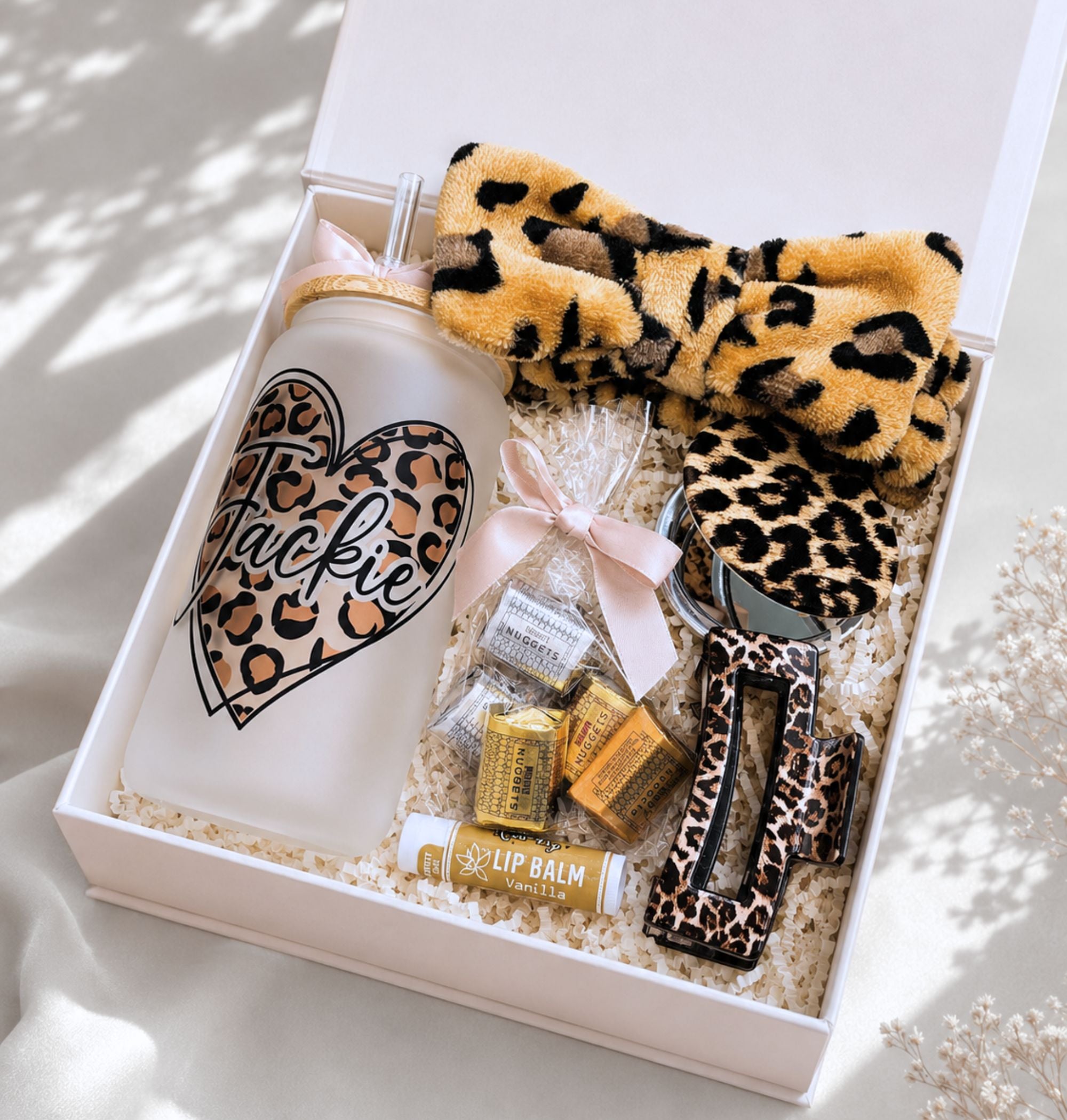 Open white magnetic gift box with leopard print self-care gift set: personalized frosted glass tumbler "Jackie" with leopard heart design, leopard spa headband, leopard compact mirror, leopard hair clip, Hershey's Nuggets in cellophane bag with pink bow, vanilla lip balm
