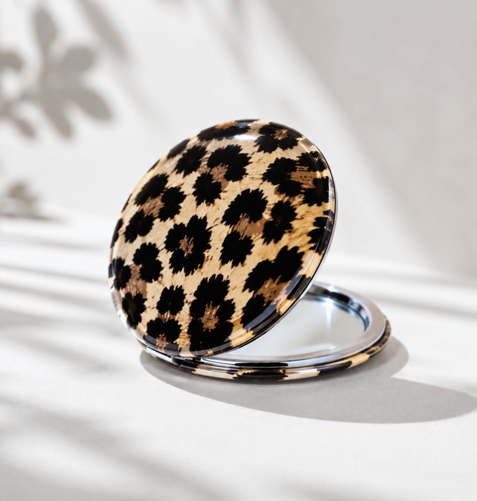 Double-sided leopard print compact travel mirror open showing silver mirror interior, styled with leaf shadow on white background