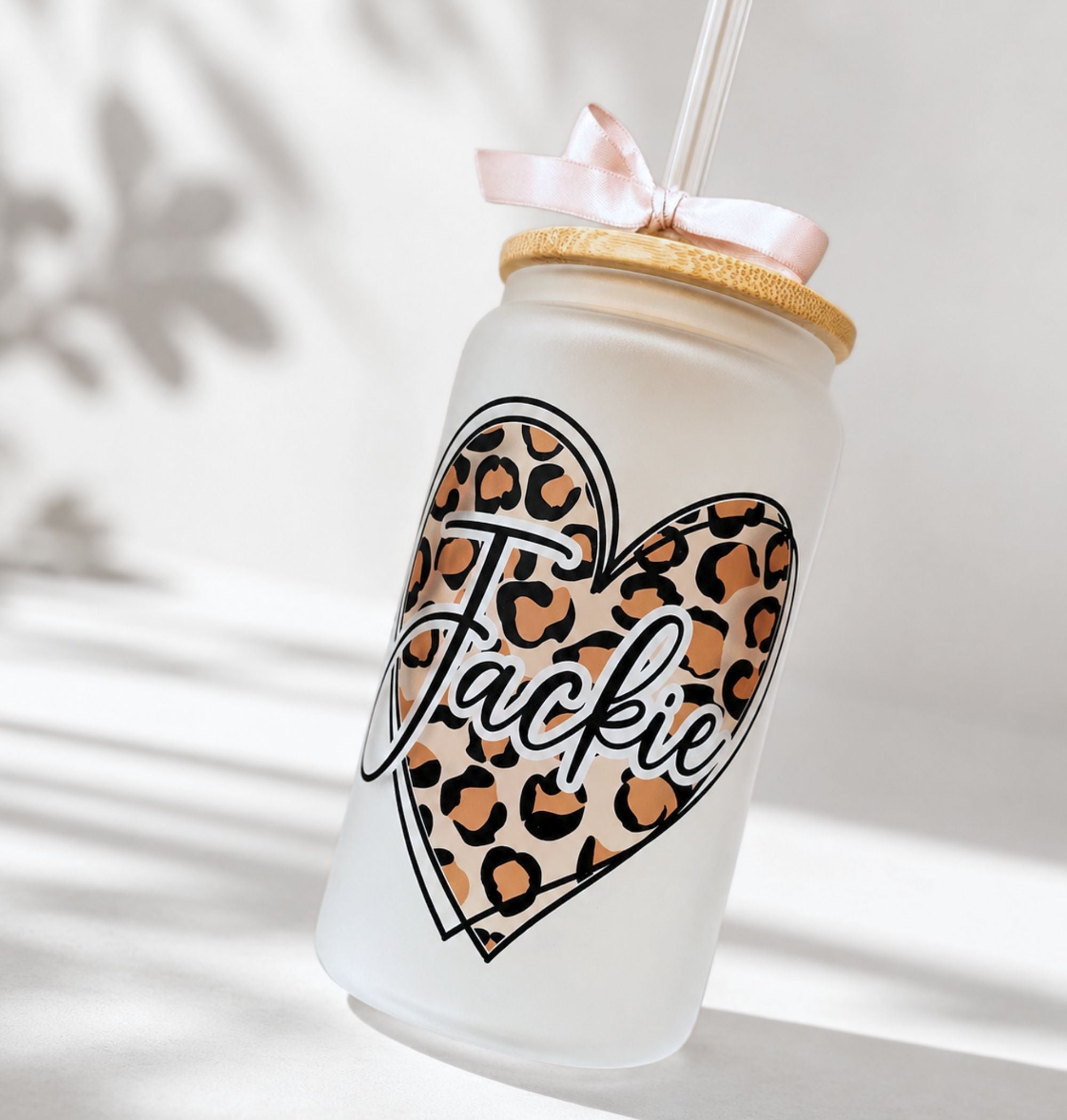 Close-up of personalized 16 oz frosted glass tumbler with leopard print heart design and "Jackie" script name, bamboo lid with pink satin ribbon bow and acrylic straw