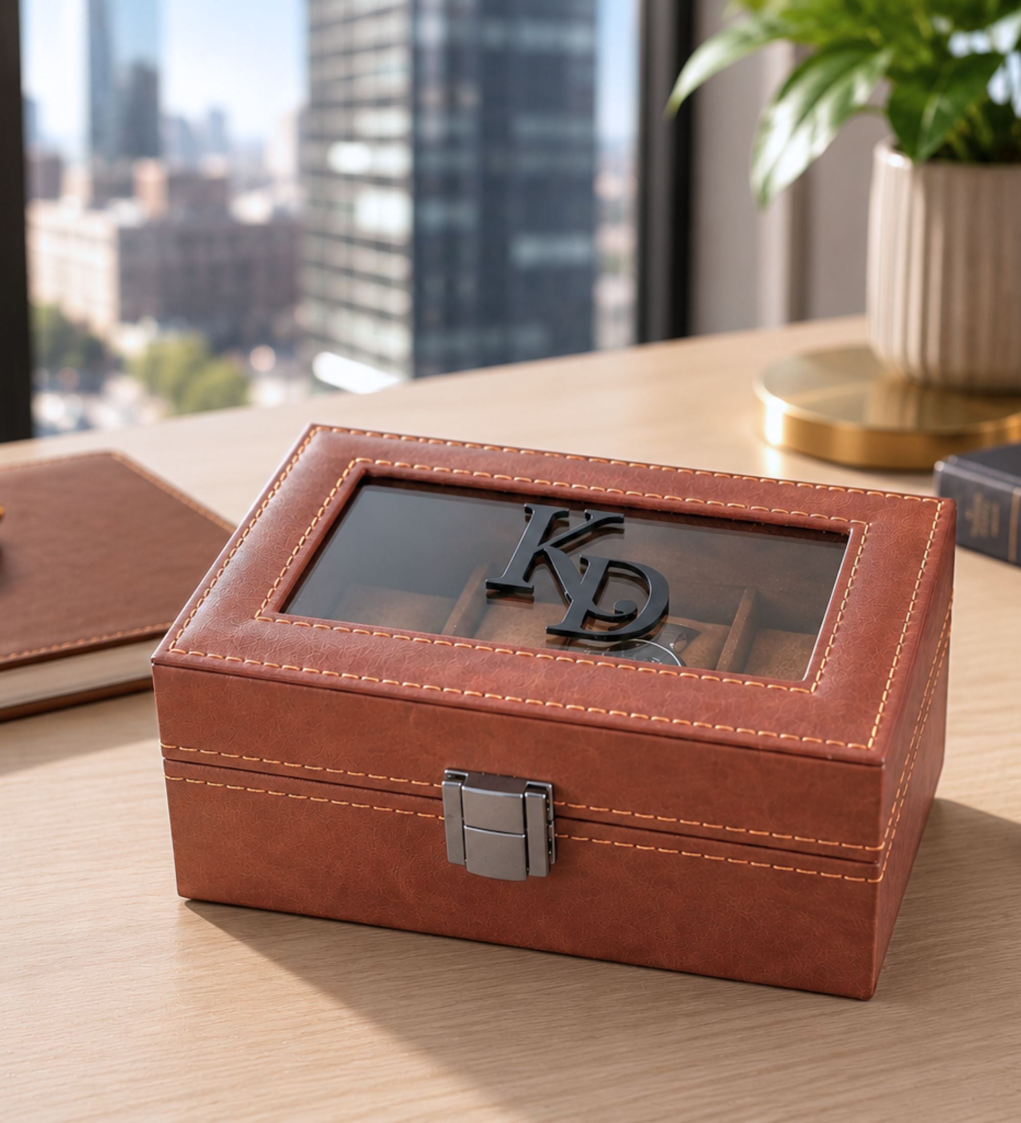 Personalized cognac leatherette watch box with black 3D acrylic monogram "KD" in Style B, displayed on a wooden desk with city skyline in background