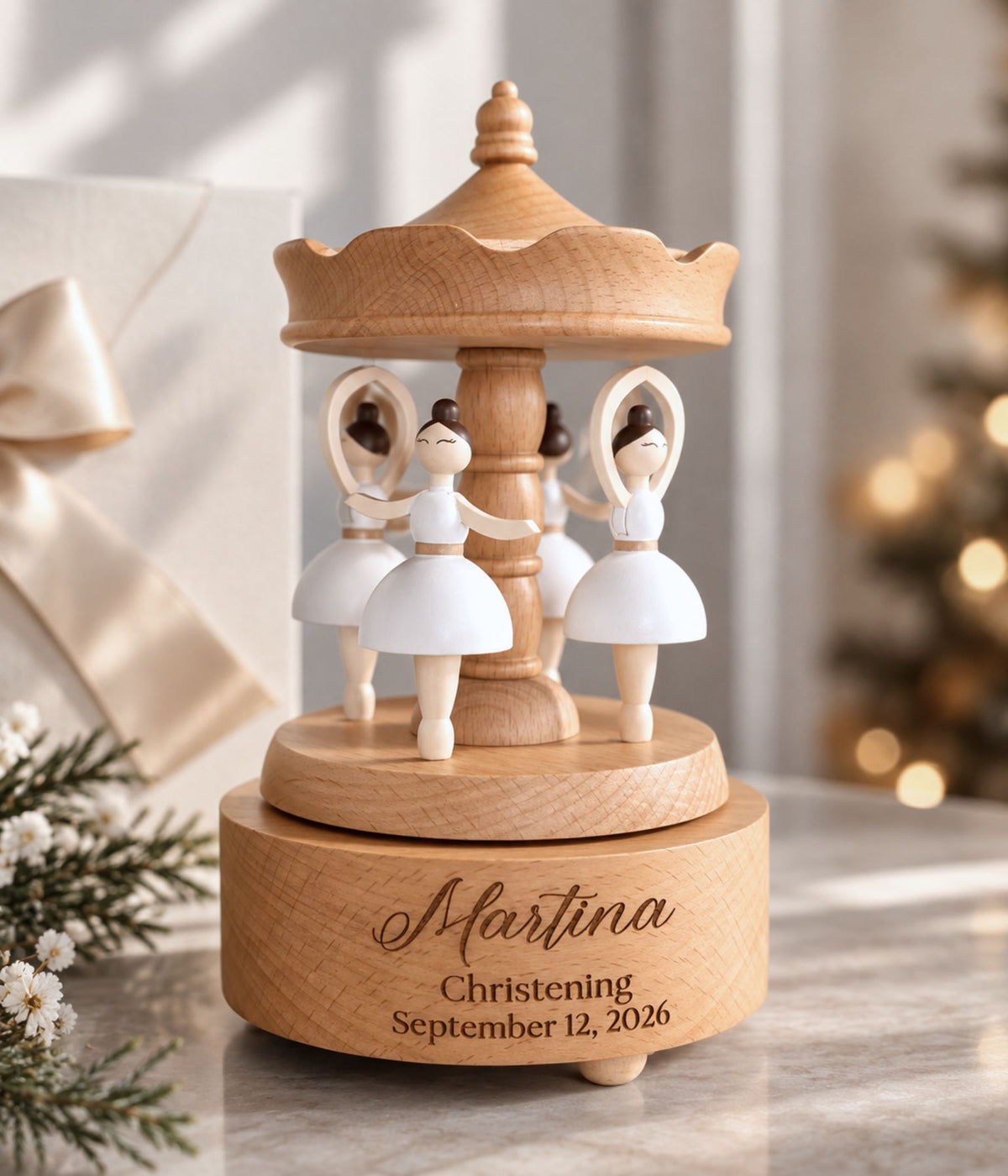 Personalized wooden carousel music box with white ballerina figures, engraved with the name 'Martina' and a christening date, styled on a marble surface with a gift box and blurred Christmas tree in the background.